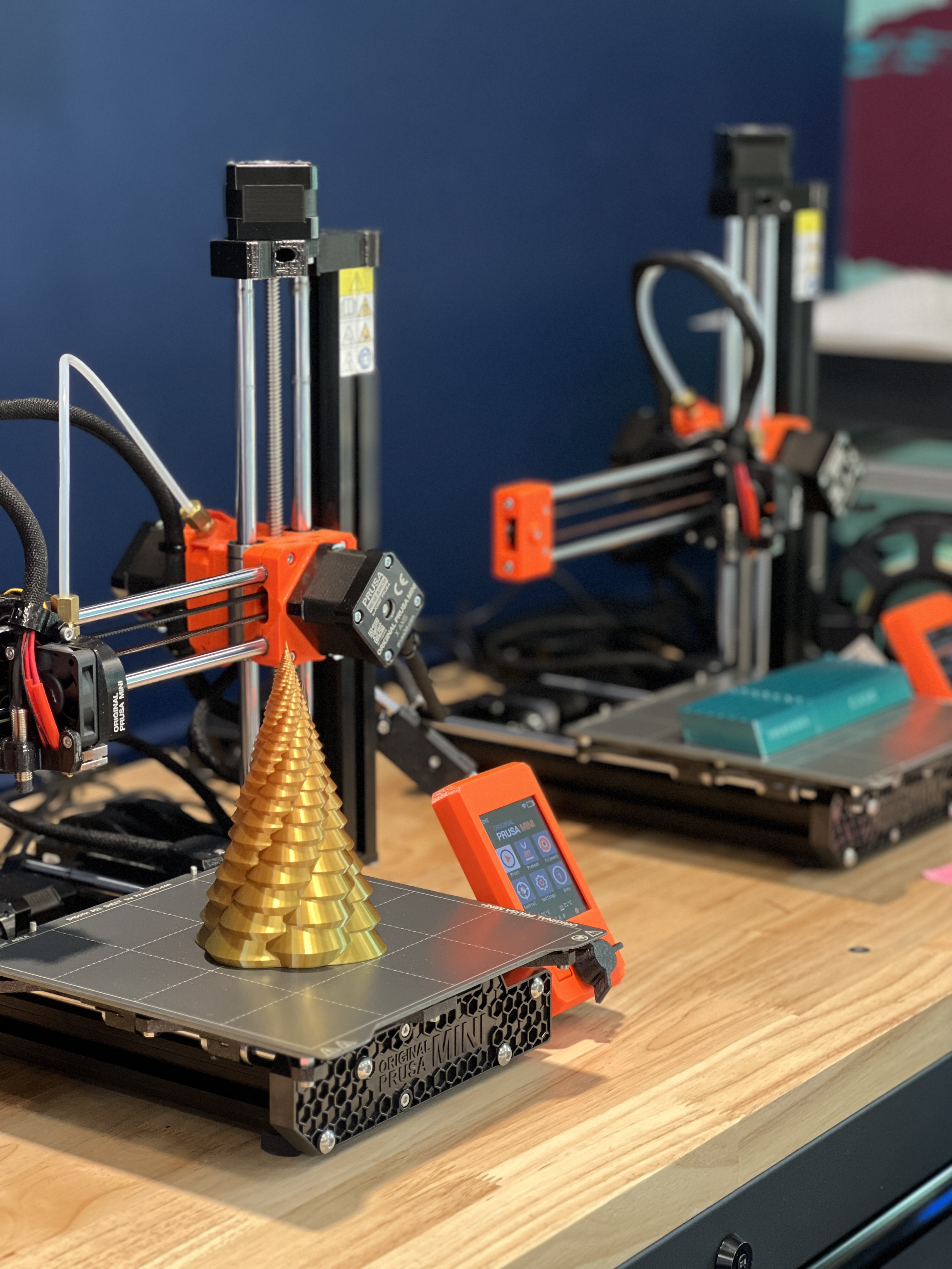 Two Prusa Mini 3D Printers. The first one has a geometric gold colored tree on the build plate.