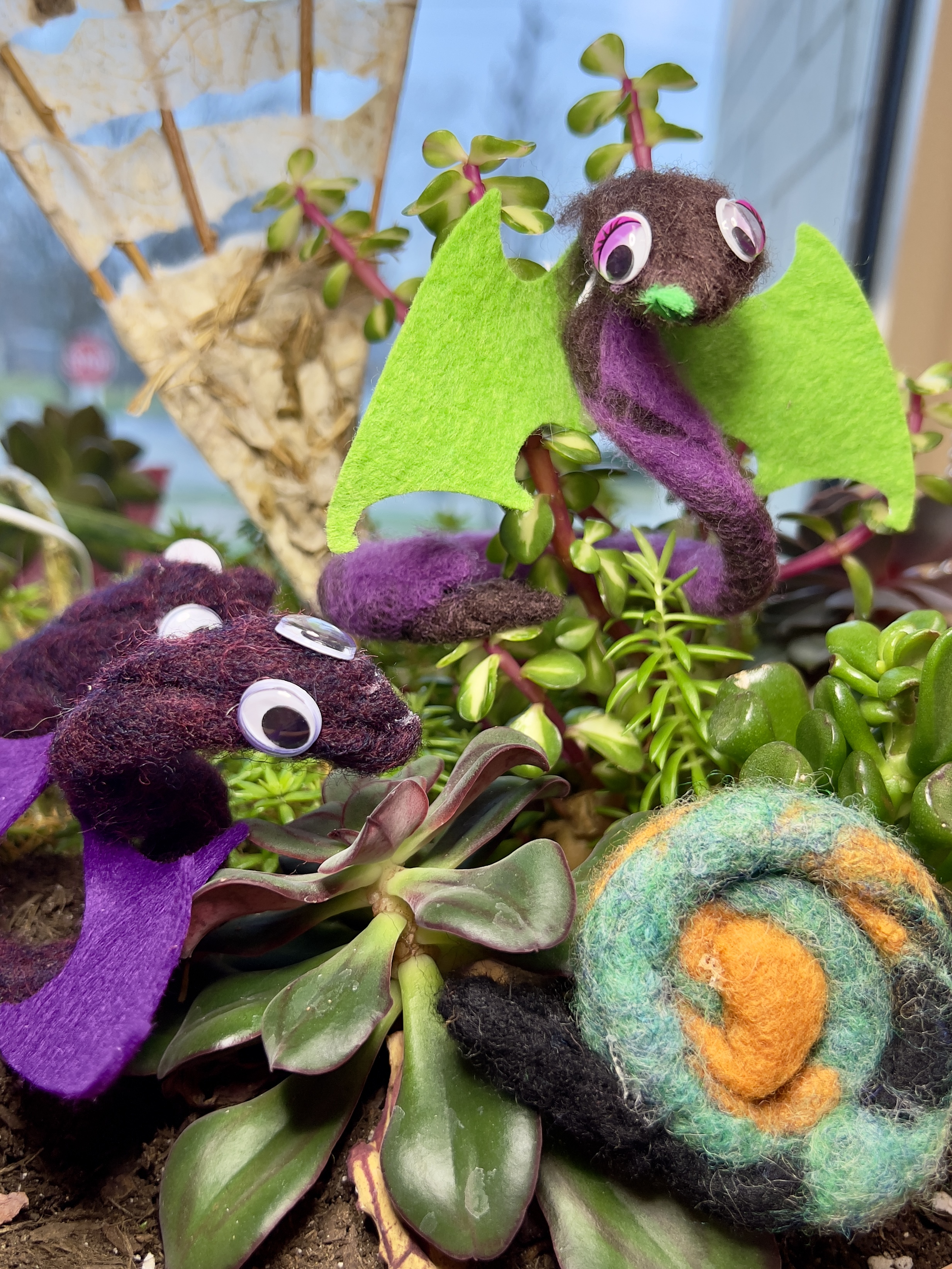 Purple felted snakes with purple and green wings and a green gold and black felted snail all sitting on plants