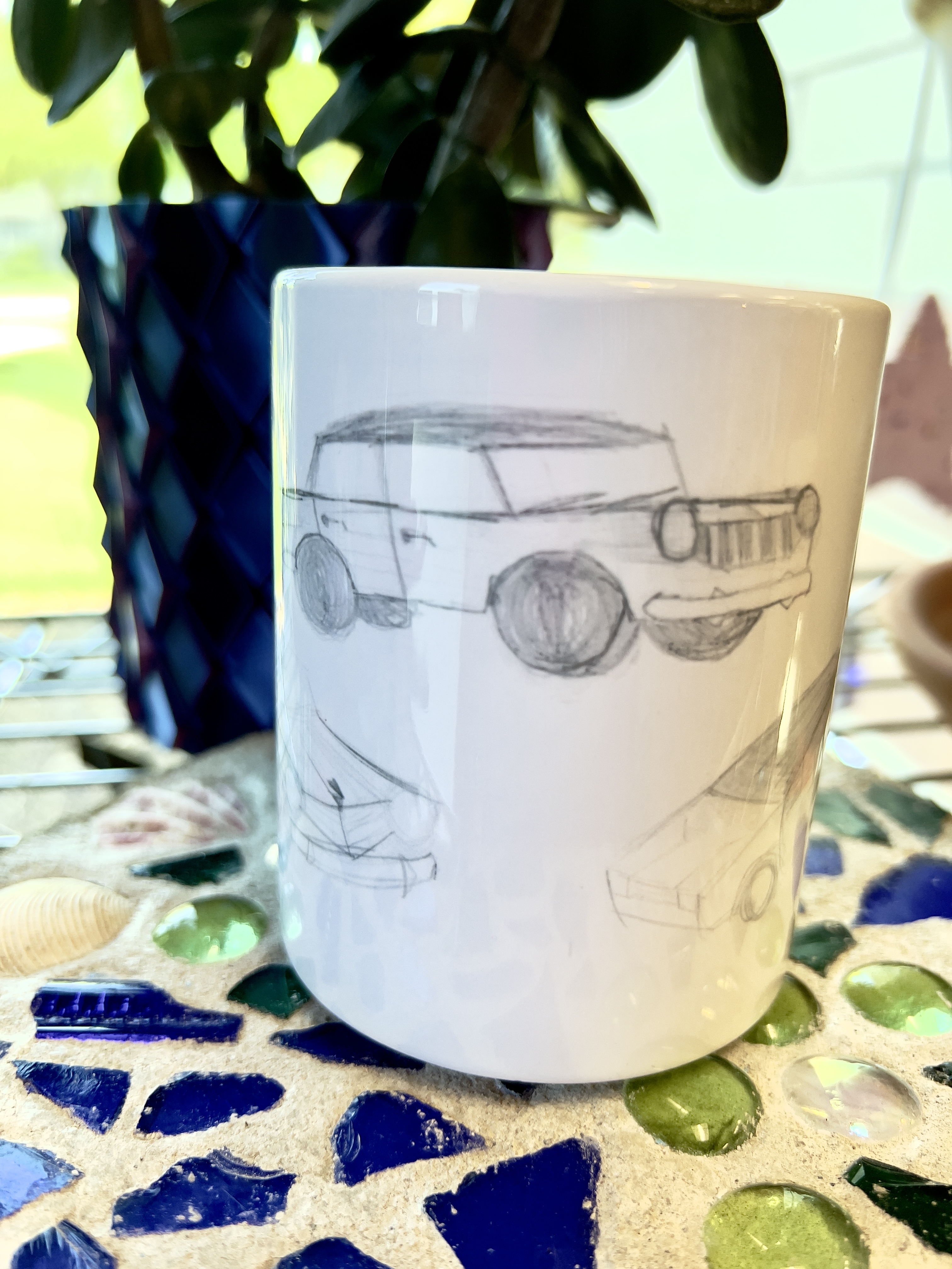 white coffee mug with cars drawn in pencil, mug is sitting on a garden stone with blue and green glass, with a plant behind mug