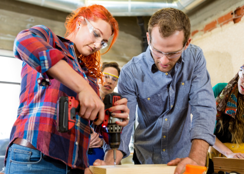 woman with red hair and power drill man holding wood to be drilled women in background watching