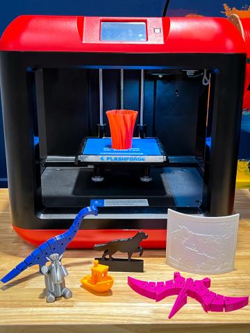 Red and black 3D printer with 3D printed dinosaurs, boat, dog, vase