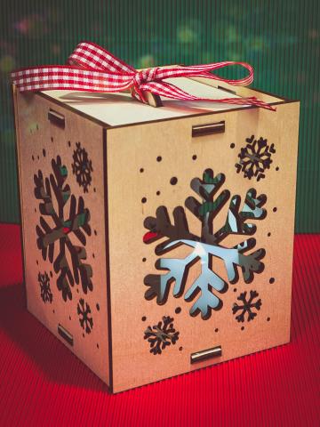 a four sided wooden laser cut box with top and bottom, laser cut snowflake designs, red and white gingham ribbon on the top, set on a red piece of cardstock with a green one for the backdrop.