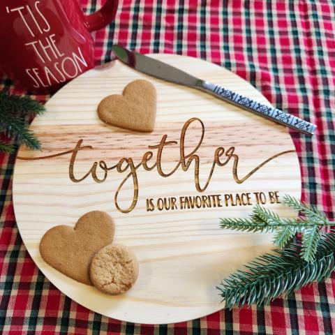 round wood piece with Together is our favorite place engraved on top with heart cookies on it and a knife and a red coffee cup, a twig of pine tree, and a Xmas gingham tablecloth.