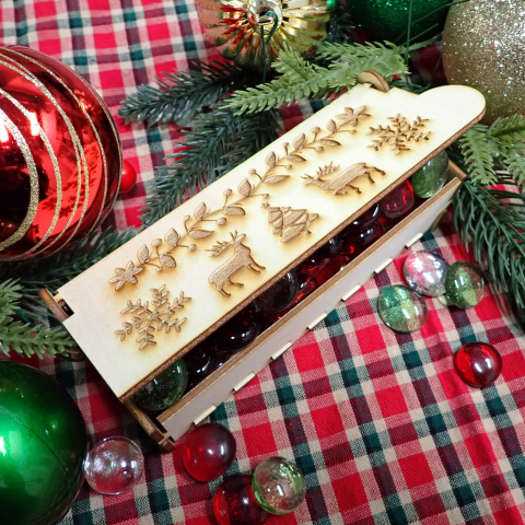 Small rectangle box made out of wood with reindeer, snowflakes, and holly and ivy engraved on top, a twig of pine tree, and red, green, and gold round ornaments, and a Xmas gingham tablecloth.