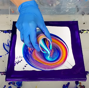 hand with blue glove and cup of multiple colors of acrylic paint being poured onto a canvas