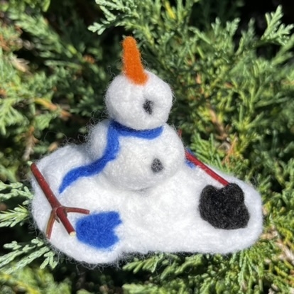 White snowman made our of wool roving. Looks like it's melting. Has a carrot nose, blue scarf, and a black hat and blue gloves.