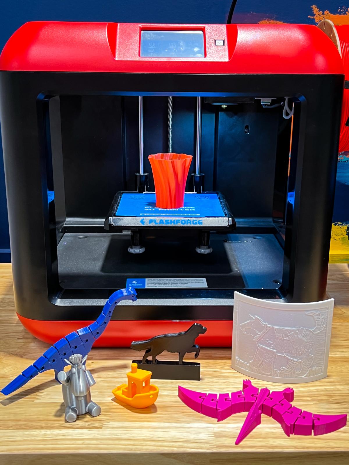 Red FlashForge Finder 3D Printer with a 3D printed vase on the print bed and 3D printed dinos, dogs, boats, and a lithophane