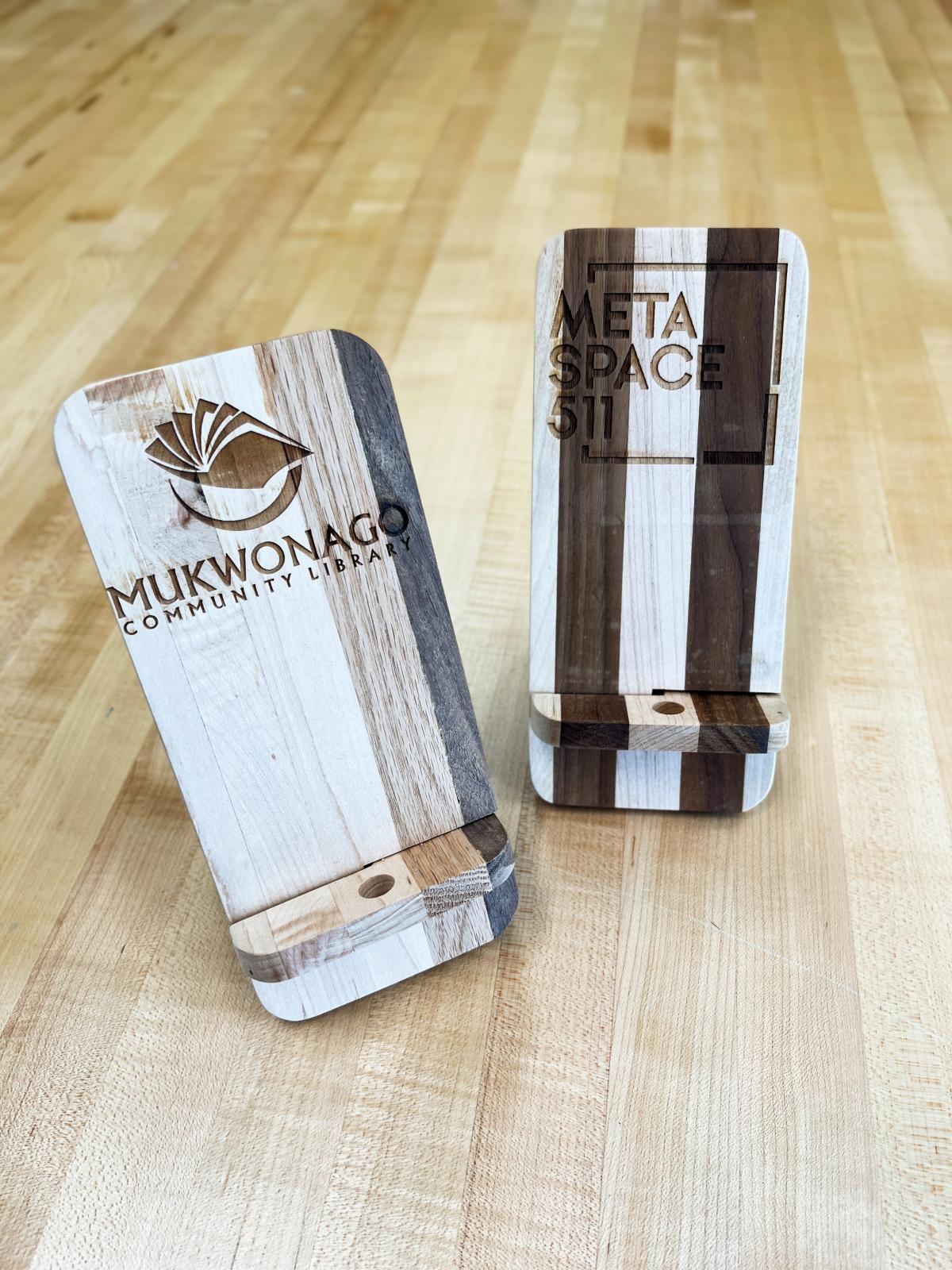 Two wooden laser engraved phone stands with the Library logo and MetaSpace logo on them.