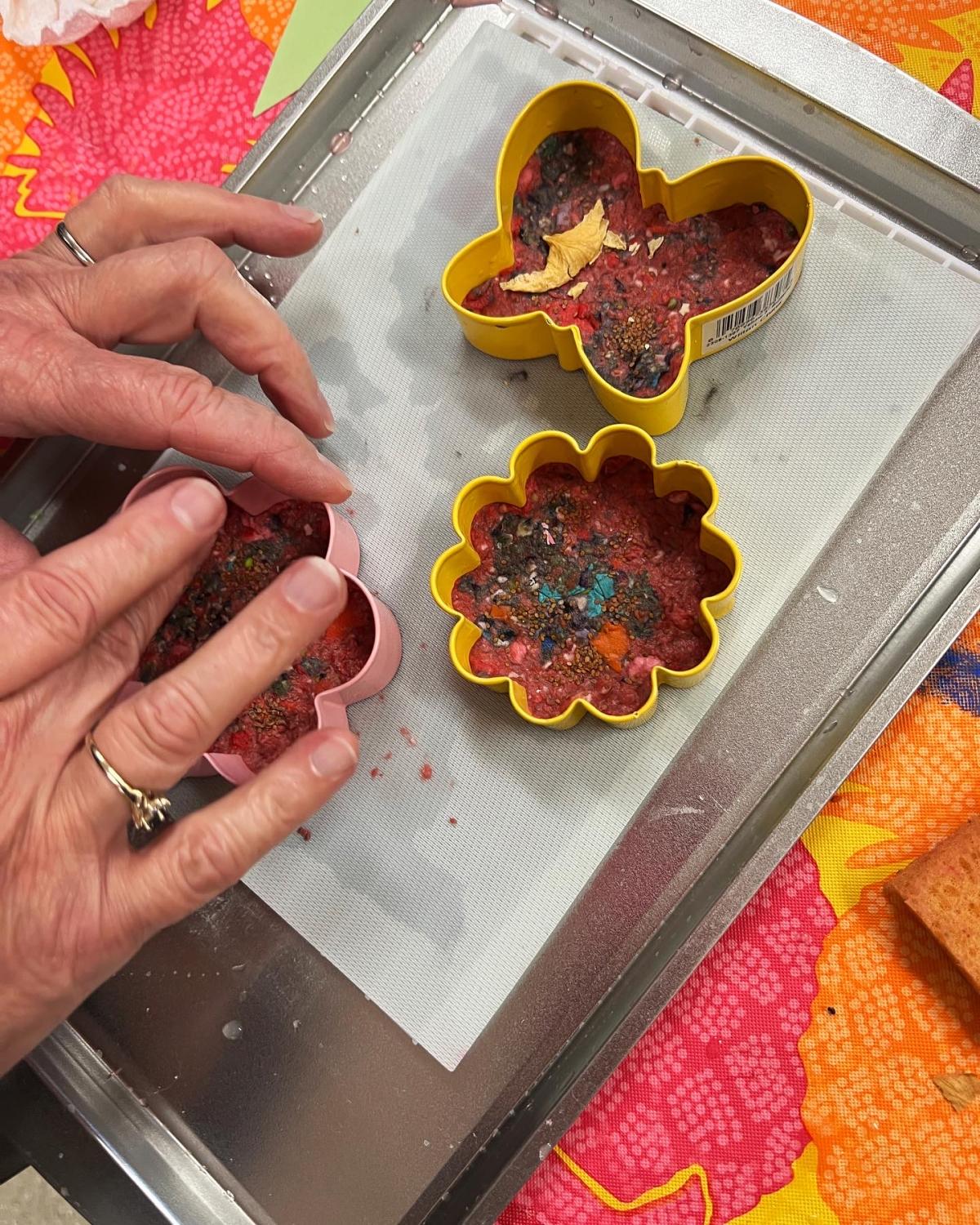 Silver cookie sheet with yellow butterfly and flower cookie cutters with red and blue mixed paper pulp in them. Hands on the left packing paper pulp into another yellow flower cookie cutter