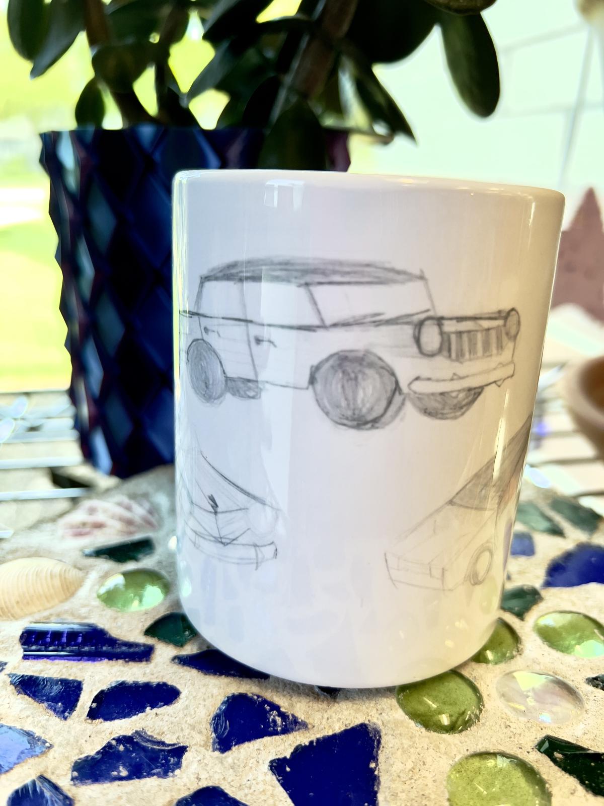 white coffee mug with cars drawn in pencil, mug is sitting on a garden stone with blue and green glass, with a plant behind mug